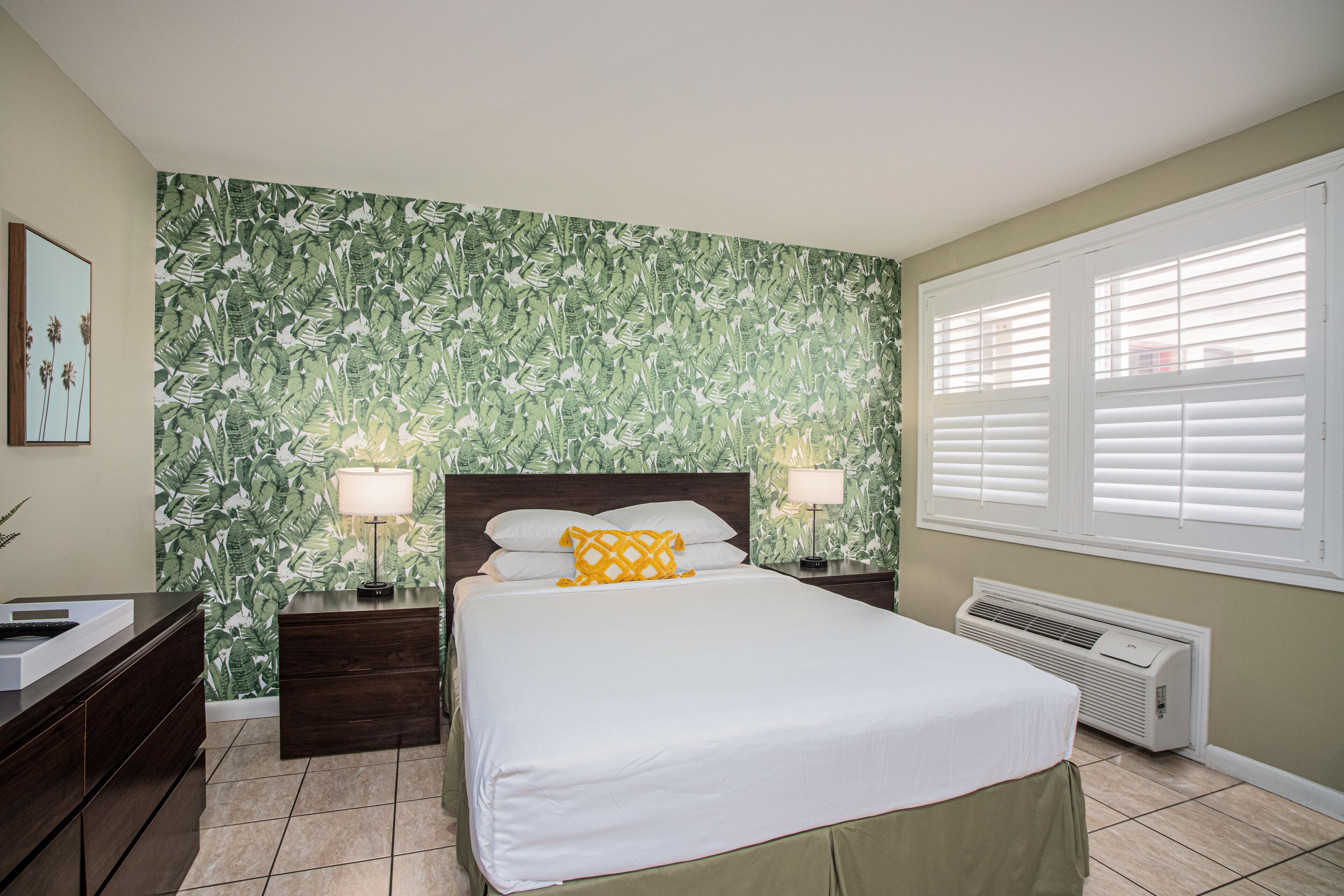 Quarto com cama, duas mesas de cabeceira com luminárias, um criado-mudo, ar-condicionado fixado na parede e uma janela com persianas.