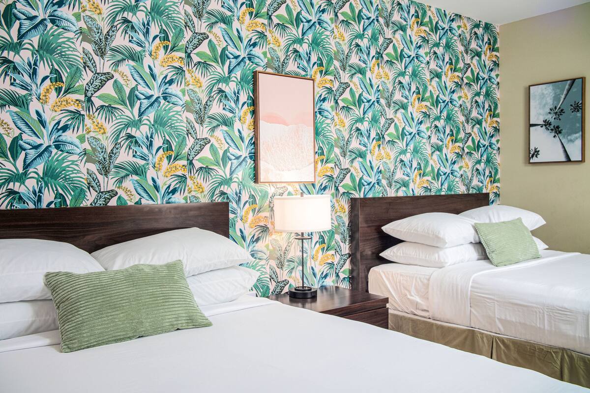 Une chambre d’hôtel avec deux lits, une table de chevet avec une lampe et un papier peint aux motifs tropicaux.