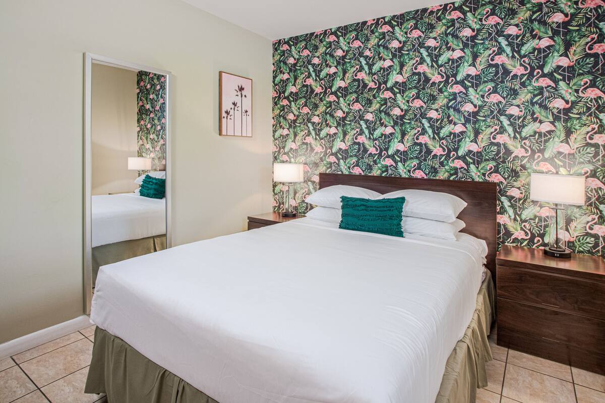 Un lit bien fait, avec du linge de lit blanc et un coussin d’accent de couleur turquoise, une tête de lit en bois et un mur recouvert d’un papier peint à motifs floraux.