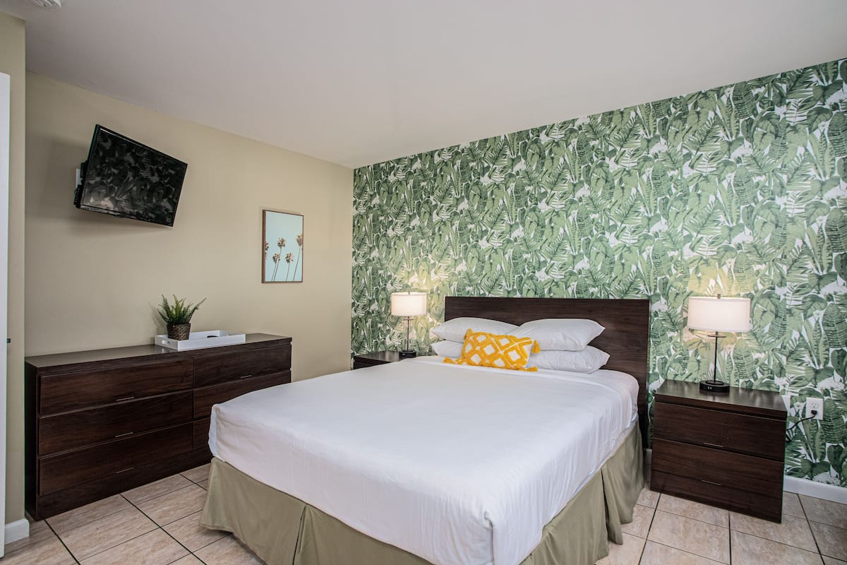 Une chambre à coucher comprenant un lit, des tables de chevet, une commode, un téléviseur, une lampe fixée au mur et une plante.
