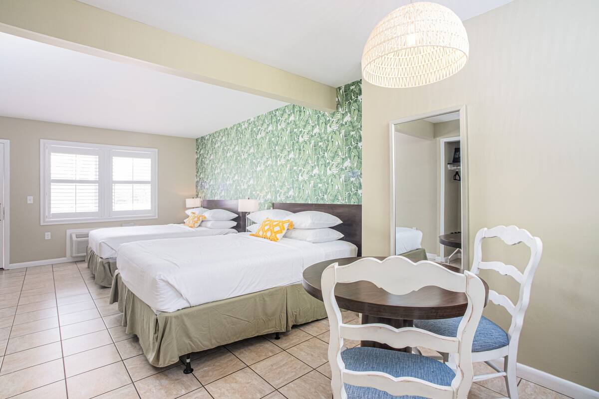 Une chambre d’hôtel avec deux lits, une table à manger avec des chaises, un luminaire suspendu et un grand miroir.