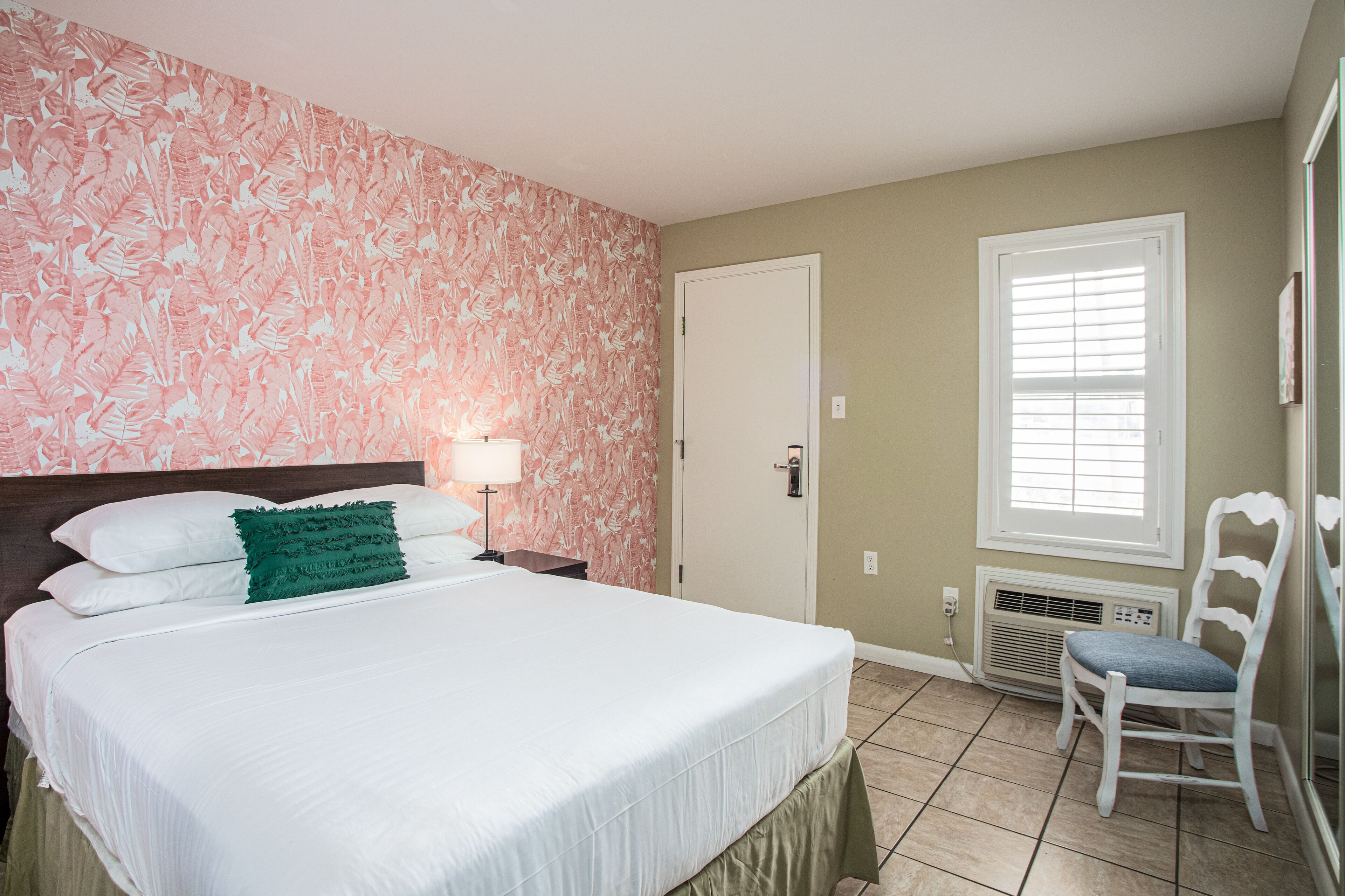 A bedroom with a bed, a chair, a nightstand, a lamp, a door, and a window with blinds.
