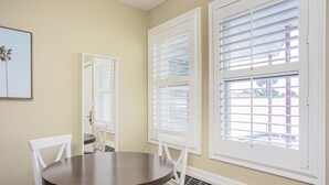 A dining area with a round table and white chairs, large windows with blinds, and a wall-mounted air vent. - The Local - St. Augustine (St. Augustine)
