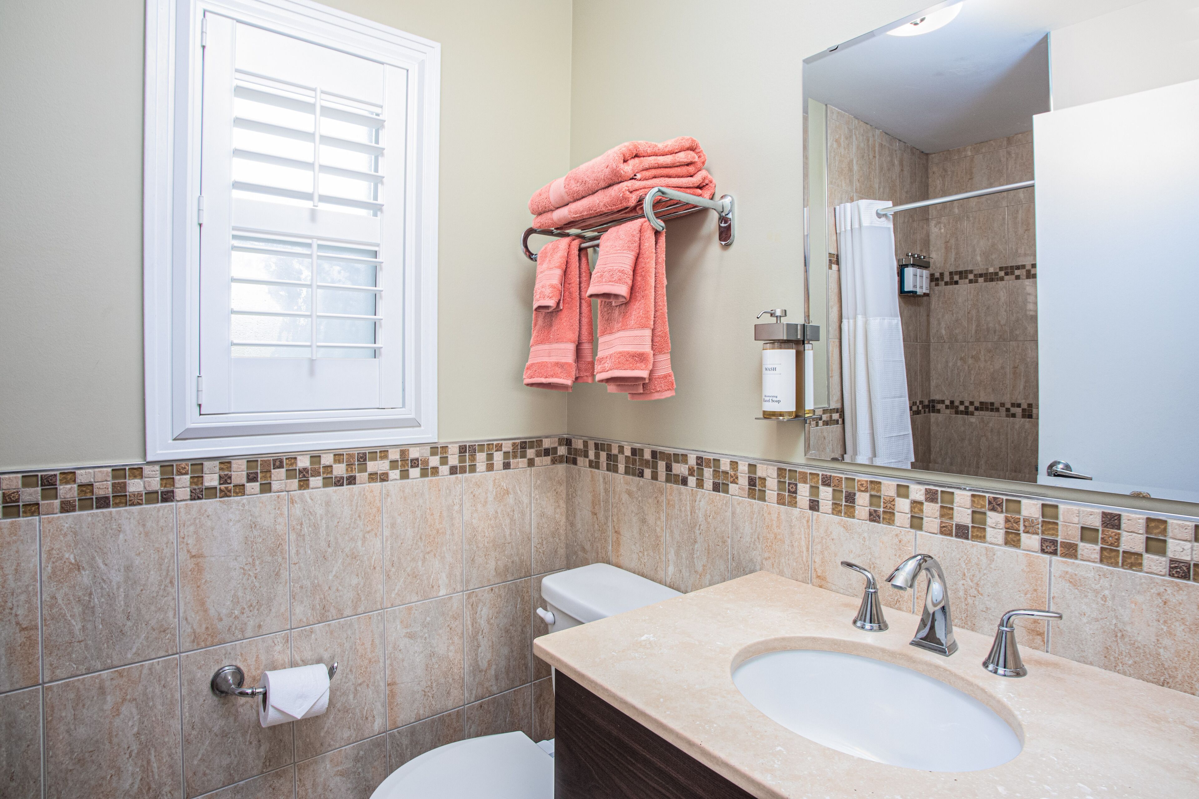 Un baño con pared de azulejos color beige, inodoro blanco, lavamanos con dos grifos y un perchero con toallas rosas.
