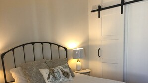 Ein ordentlich bezogenes Bett mit weißer Daunendecke, einem Metallkopfteil und gemusterten Kissen. Ein Nachttisch mit Lampe und einem kleinen dekorativen Gegenstand ist vorhanden. Der Raum verfügt über Holzboden und eine weiße Tür mit schwarzem Griff.