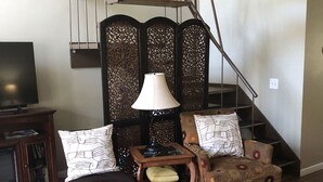 Ein Wohnzimmer mit Holzboden, einer Couch, einem Sessel, einem kleinen Tisch mit einer Lampe und einem Fernseher. Eine Treppe mit Geländer und einem dekorativen Raumteiler ist ebenfalls zu sehen.