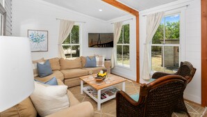 Un salón amplio con un sofá color crema, una mesa de centro de madera y sillas de mimbre. Hay ventanas grandes con cortinas, un televisor de pantalla plana montado en la pared y un cuadro enmarcado.