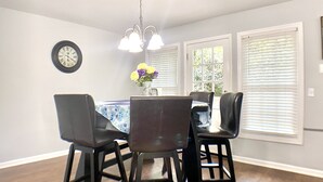 Uma área de jantar com uma mesa, cadeiras, um relógio e uma janela com persianas.