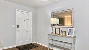 Um corredor com porta branca, piso de madeira, mesa consola com espelho e quadros emoldurados.