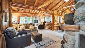 Interior de una cabaña de troncos con techo de madera, chimenea, sofá y televisor.