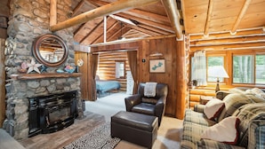 Interior de una cabaña de troncos con chimenea de piedra, sillón de cuero, sofá escocés y un ventanal con vistas al exterior.
