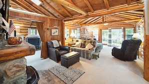 Interior acogedor de una cabaña de madera con chimenea, asientos de cuero y ventanas amplias que ofrecen vistas al exterior.