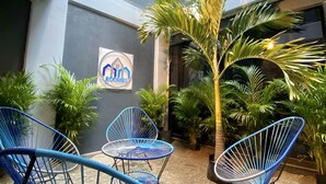 Een binnentuin met blauwe draadstoelen, planten in potten en een bord met de tekst 'ROOMS'.