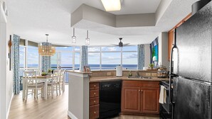 Une cuisine moderne avec vue sur l’océan, des meubles en bois, un réfrigérateur noir et un coin repas avec une table et des chaises.