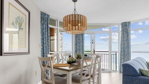 Un coin repas avec une table et des chaises en bois, un luminaire suspendu en osier, et une vue sur l’océan grâce à de grandes fenêtres.