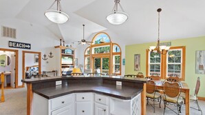 A kitchen with a central island, wooden cabinets, and a dining area with wicker chairs. - Castaway: Private Pool, Hot Tub & Ocean Breeze (Corolla)
