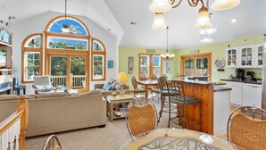 A spacious living room with a large window, a dining area with a bar, and a kitchen with white cabinets. - Castaway: Private Pool, Hot Tub & Ocean Breeze (Corolla)