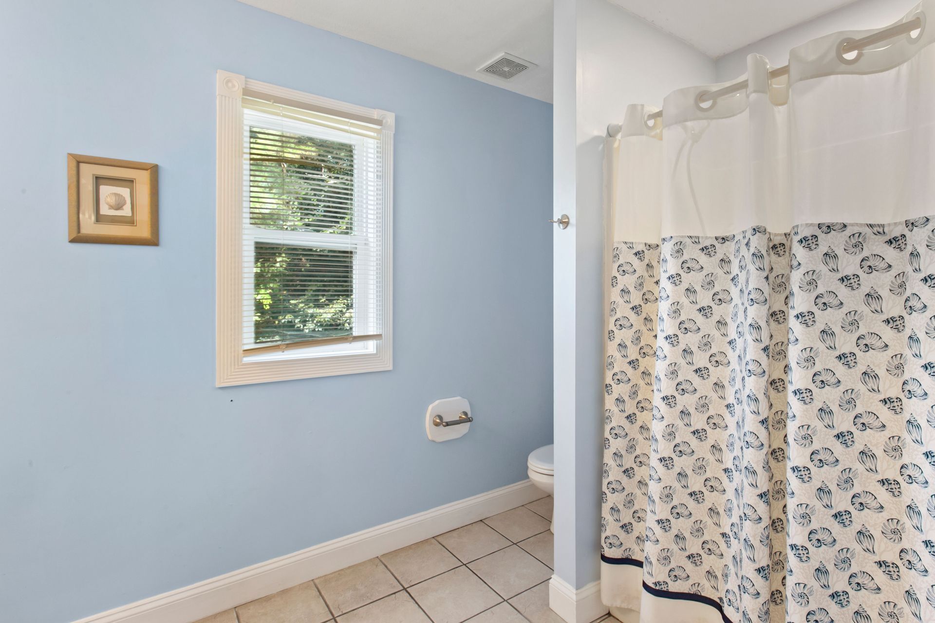 A bathroom with a window, toilet, and shower curtain with a shell pattern.