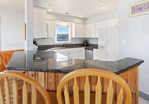 A kitchen with a granite countertop, wooden chairs, white cabinets, and a window.
