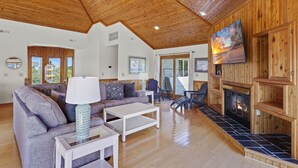 A spacious living room with a wooden ceiling, a fireplace, and a large TV mounted on the wall. - Nautica Nook: Ocean Escape with Saltwater Pool (Corolla)