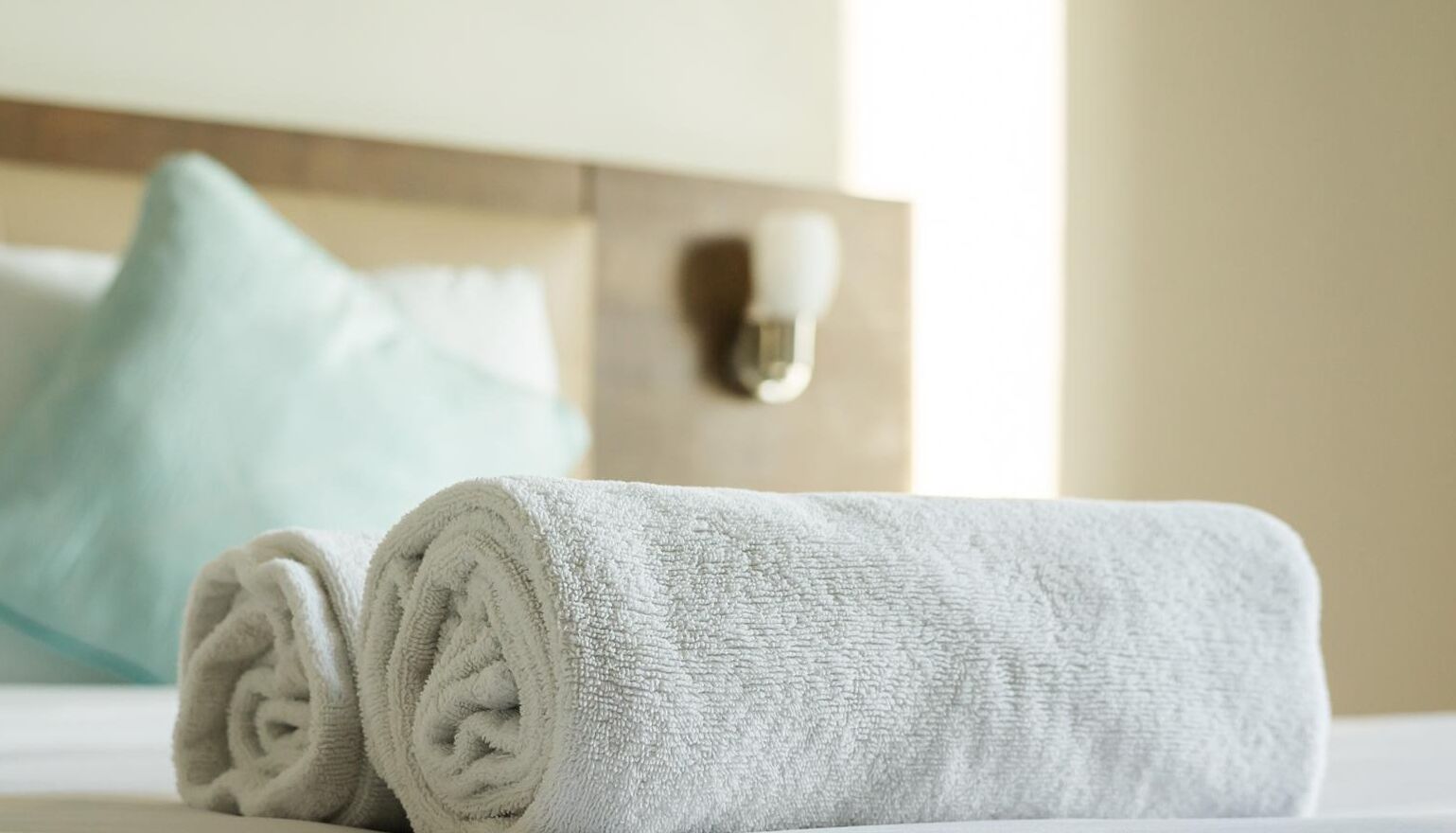 Un asciugamano bianco e ordinato su un letto con una testiera in legno e un cuscino azzurro sullo sfondo.