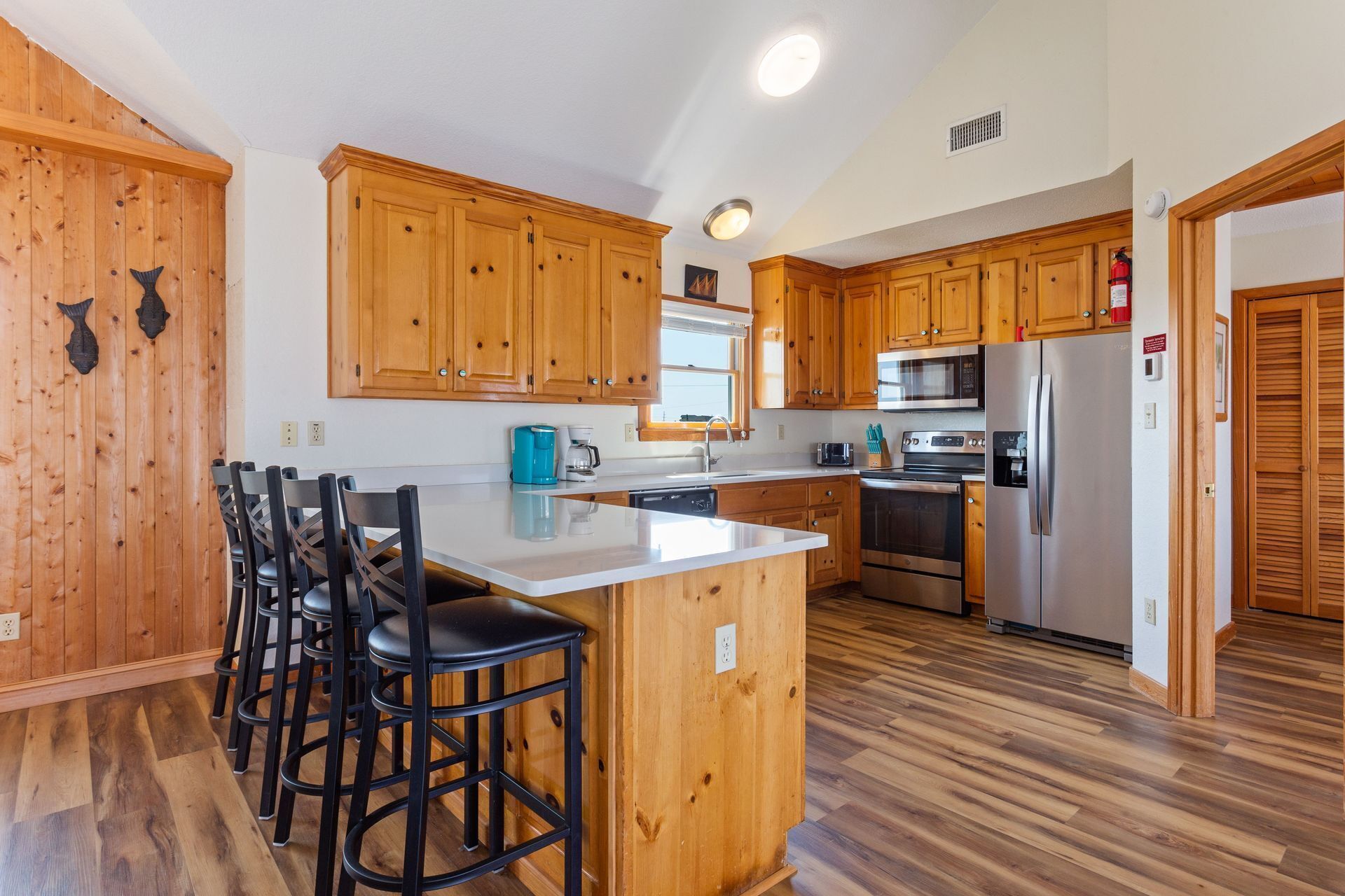 A kitchen with wooden cabinets, a stainless steel refrigerator, a microwave, and a sink. There are bar stools and a window above the sink.