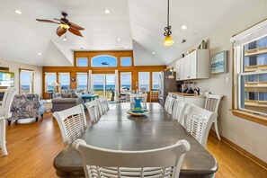 A spacious dining area with a large table, white chairs, and a view of the ocean through large windows. - 7000 Serenity Sands*Pool*Dog Friendly*Pool Table (Nags Head)