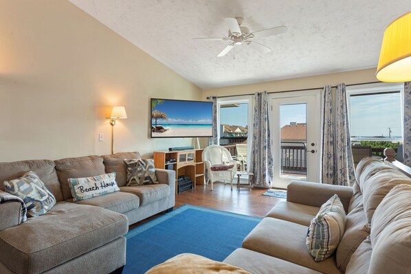 Ein Wohnzimmer mit einer Sofagarnitur, einem Fernseher, einem Deckenventilator und Blick auf den Strand durch die geöffnete Tür.