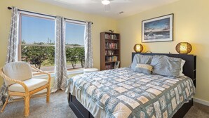 A bedroom with a bed, a chair, a bookshelf, and a window with curtains. - Pet-Friendly Sea Dunes Townhome Near Beach (Kitty Hawk)
