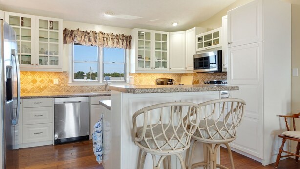 A kitchen with white cabinets, a stainless steel refrigerator, a microwave, and a coffee maker. There is a window with a view of the outdoors, and two wicker chairs at the island. - Pet-Friendly Sea Dunes Townhome Near Beach (Kitty Hawk)