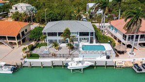 Une grande maison avec une piscine, un quai avec un bateau, et un petit bateau amarré au ponton.