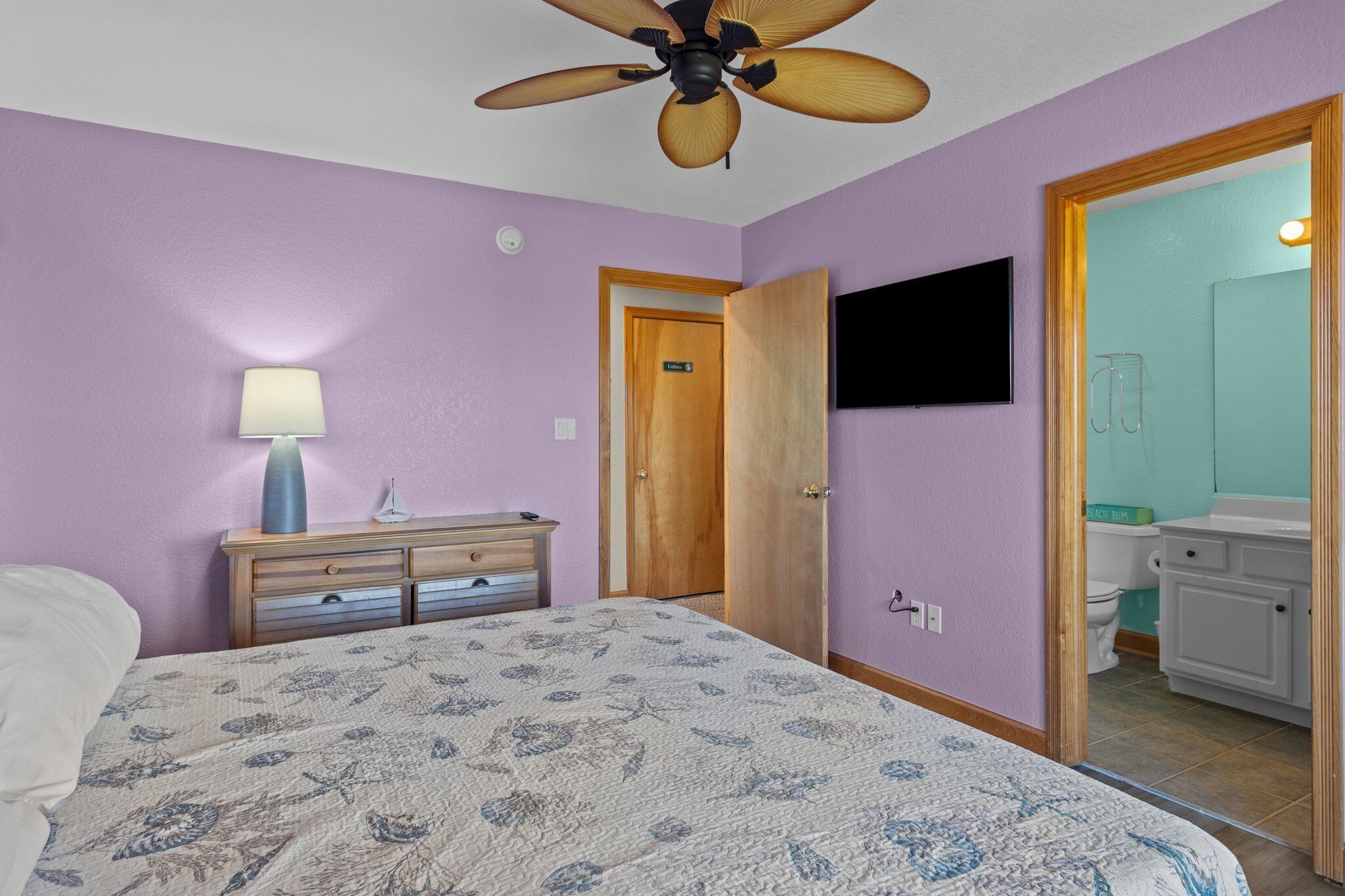 A bedroom with a bed, a ceiling fan, a dresser, a TV, and a bathroom visible through an open door.
