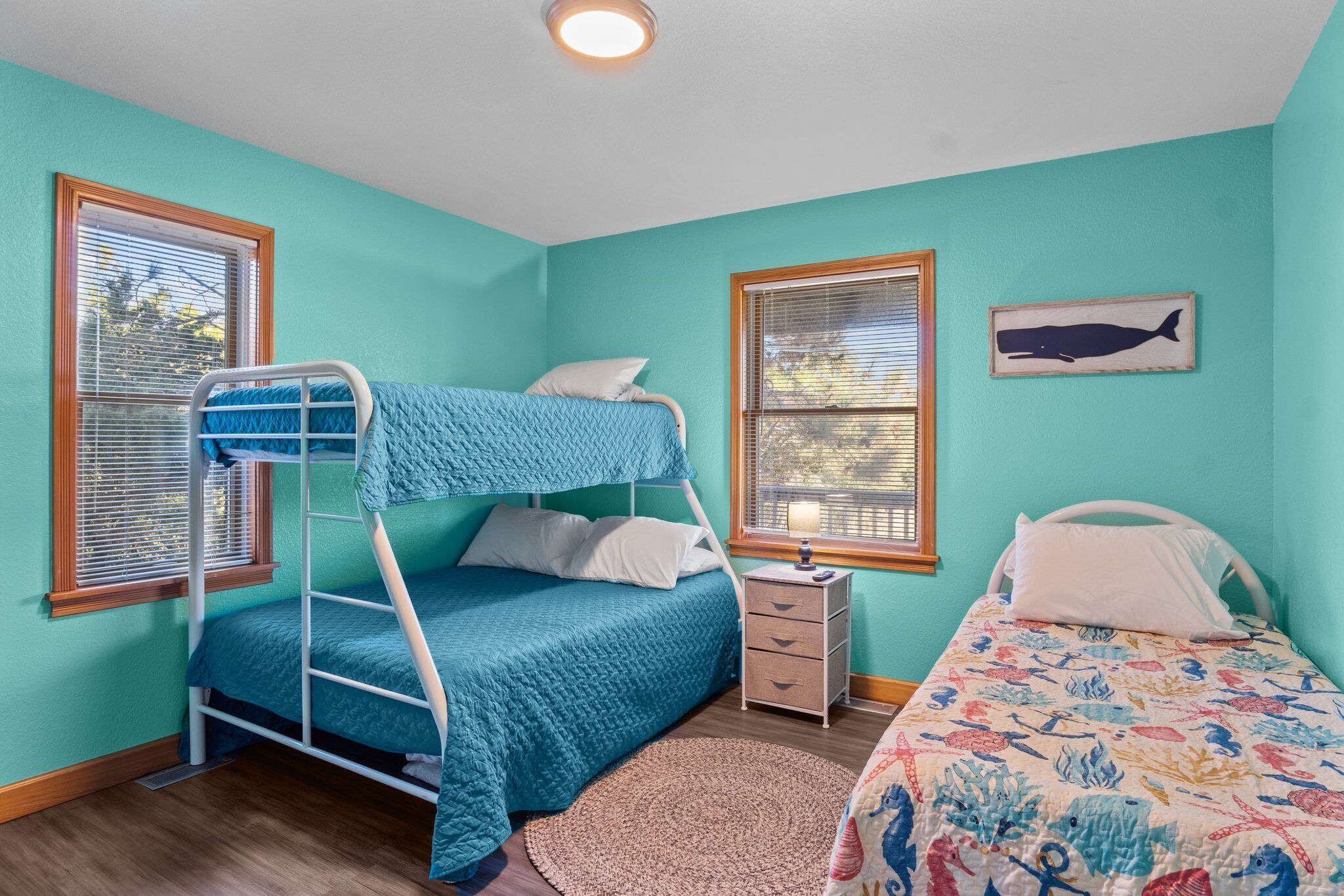A bunk bed room with teal walls, a window with blinds, a nightstand, and a bed with a colorful quilt.