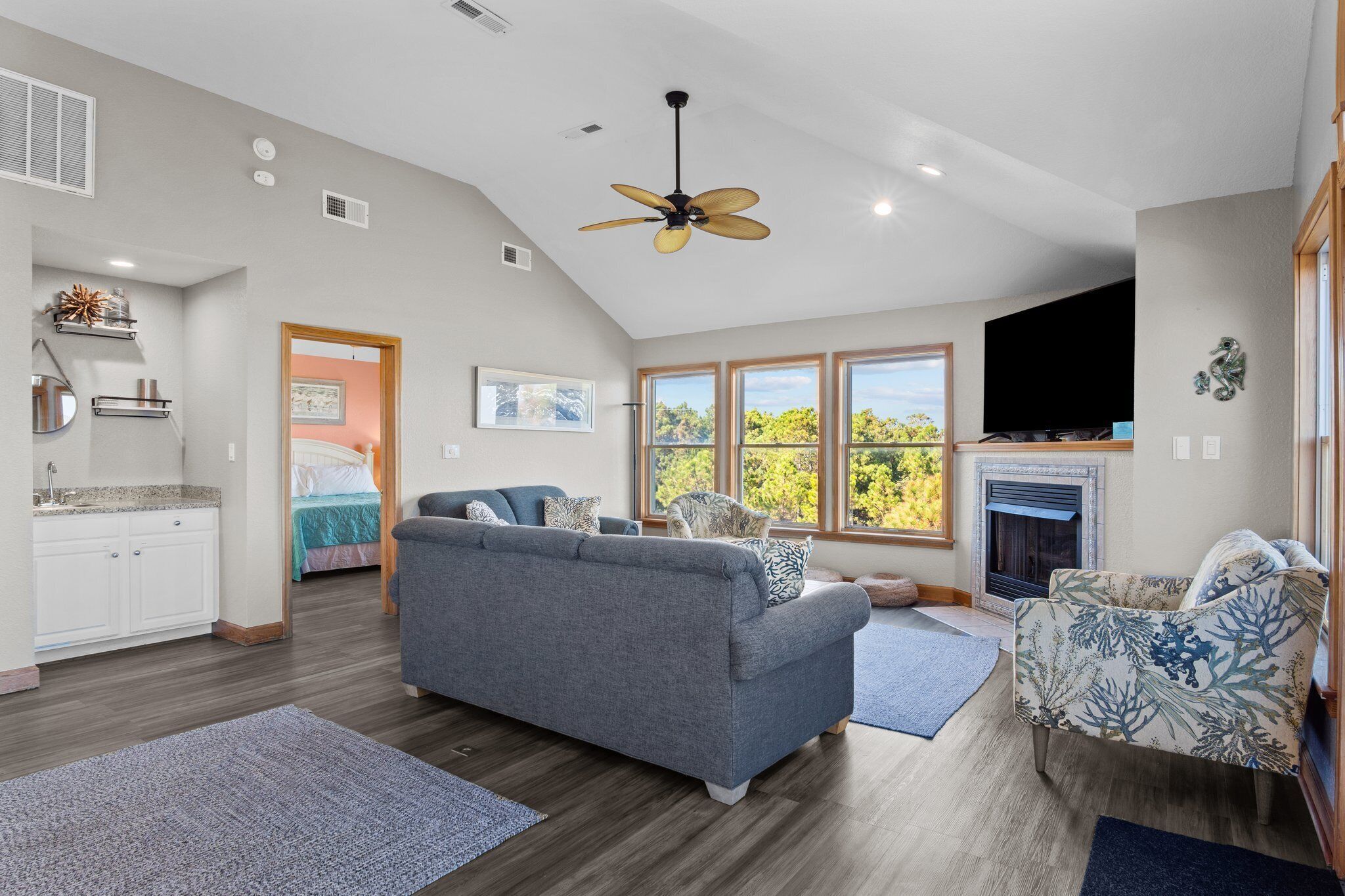A spacious living area with a grey sofa, a patterned armchair, a fireplace, and large windows offering a view of trees.