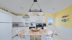Una cocina con armarios blancos, una mesa de comedor de madera y sillas blancas. Hay un microondas, una cafetera y una cesta en la mesa.