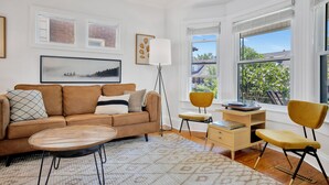 A modern living room with a brown sofa, a coffee table, a desk with a chair, and large windows with a view of greenery. - The Hipster House  (Windsor)