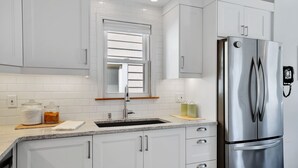 A modern kitchen with white cabinets, a stainless steel refrigerator, a window with blinds, and a granite countertop. - The Hipster House  (Windsor)