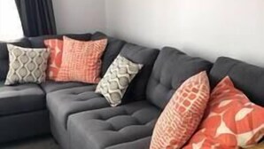 Un canapé d’angle gris avec des coussins à motifs, dans une pièce dont un mur est peint en blanc et où se trouve un rideau foncé.