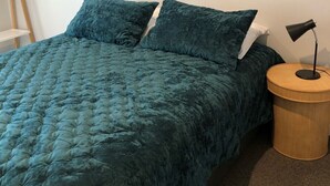 Un lit recouvert d’une courtepointe turquoise à motifs matelassés, avec deux oreillers, une table de chevet ronde en bois surmontée d’une lampe noire, et un mur blanc contre lequel est appuyée une échelle en bois.