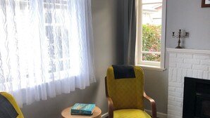 Un salon avec deux fauteuils jaunes, une petite table ronde sur laquelle sont posés des livres, une cheminée blanche et une fenêtre ornée de rideaux légers.
