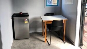Een kleine kamer met een enkel tafeltje en stoel, een kleine koelkast, en een raam met uitzicht op buiten.
