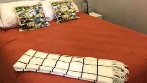 Een slaapkamer met een rode sprei, twee kussens met een collage ontwerp, en een witte deken met een ruitpatroon.