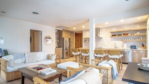 A modern living room with a sofa, chairs, and a coffee table. There is a kitchen area with a refrigerator, stove, and bar stools.