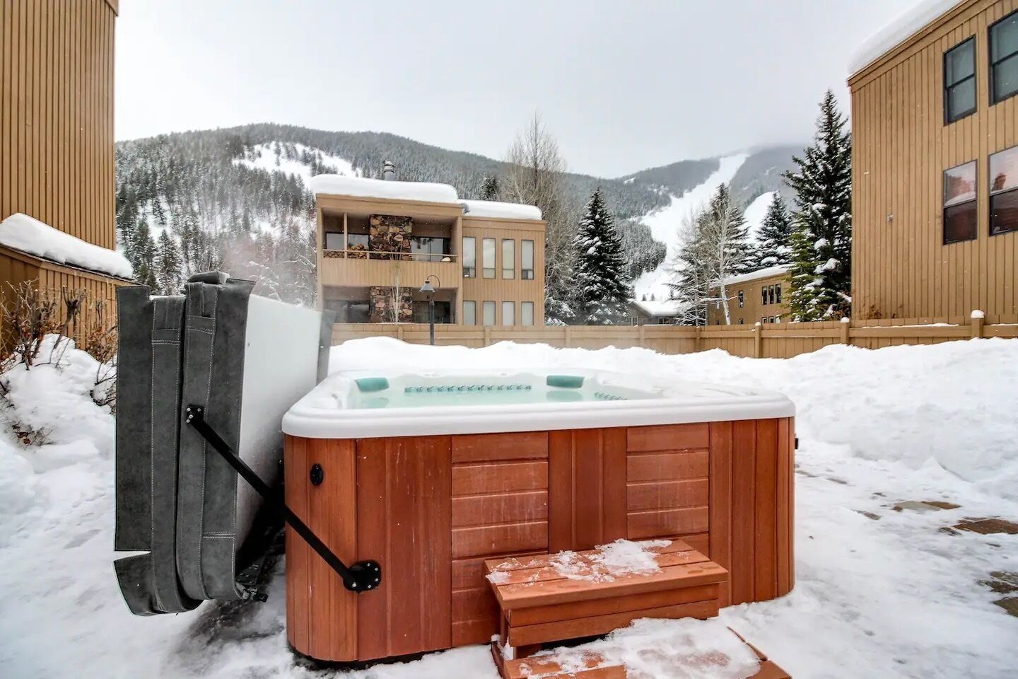 Un jacuzzi de madera con un banco delantero, situado en un espacio exterior nevado con montañas al fondo.