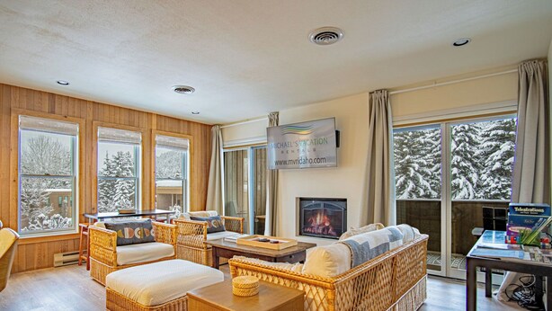 A cozy living room with wicker furniture, a fireplace, and large windows offering a snowy view.
