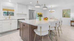 A modern kitchen with white cabinets, a large island, pendant lighting, and a painting on the wall. - Yacht Club - Walk to Beach, Restaurant/Bar! Heated Saltwater Pool  - Villa Beachside Bliss - Roelens (Cape Coral)