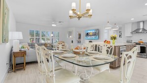A modern kitchen with a dining area, featuring a glass-top table, white chairs, and a chandelier. - Yacht Club - Walk to Beach, Restaurant/Bar! Heated Saltwater Pool  - Villa Beachside Bliss - Roelens (Cape Coral)
