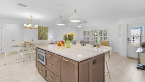 A modern kitchen with a central island, white cabinetry, and a dining area with a white table and chairs. - Yacht Club - Walk to Beach, Restaurant/Bar! Heated Saltwater Pool  - Villa Beachside Bliss - Roelens (Cape Coral)