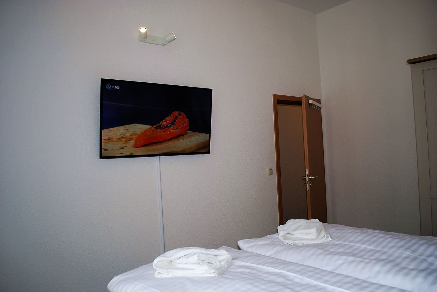 Una habitación de hotel con una cama, una toalla doblada, un televisor montado en la pared y una puerta.