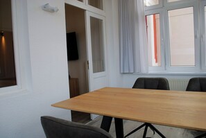 A room with a wooden table and chairs, a television in the corner, and a window with curtains. - Villy Emmy Appartement 01 A - Villa Emmy (Heringsdorf)